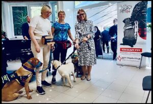 Na zdjęciu Prezeski Dogs For Life z Panią Dyrektor Ośtodka w Laskach każda z Pań ma na smyczy psa stoją w sali ośrodka